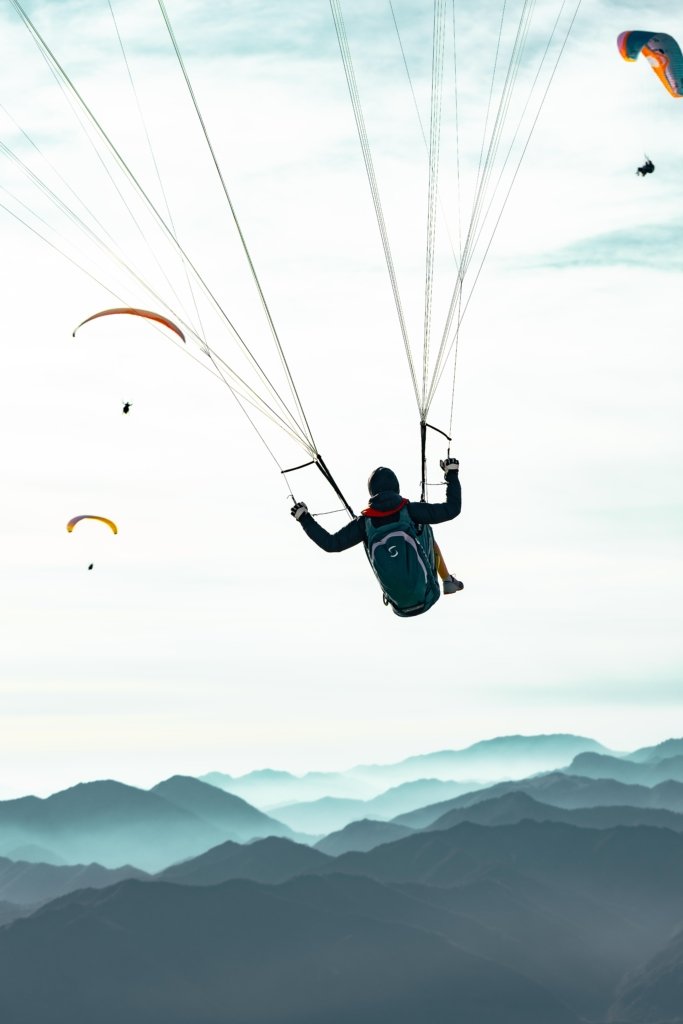 Paraglider von hinten fotografiert hoch über den blau-grauen Bergschichten am Gardasee, weitere Paraglider im Hintergrund sichtbar, atmosphärische Dunstschichten zwischen den Berggipfeln.
