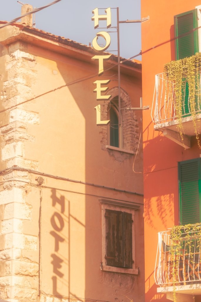 Vertikales Hotel-Schild mit goldfarbenen Buchstaben an einer orangefarbenen Hausfassade am Gardasee, dramatischer Schattenwurf des Schildes auf der angrenzenden Steinmauer, grüne Fensterläden sichtbar.