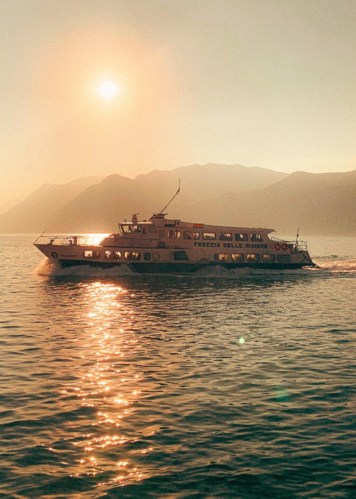 Fähre Freccia delle Riviere auf dem Gardasee bei Sonnenuntergang, goldene Sonnereflexion auf dem Wasser, Bergsilhouetten im Dunst im Hintergrund.