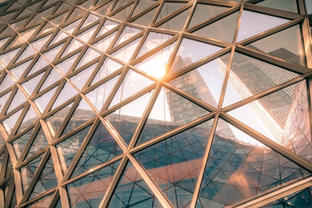 Geometrische Glasfassade der MyZeil Frankfurt mit dreieckigen Glaspaneelen, Sonnenreflektion als Lichtstrahl im Zentrum, Frankfurter Hochhäuser im Glas gespiegelt, warmes goldenes Licht.