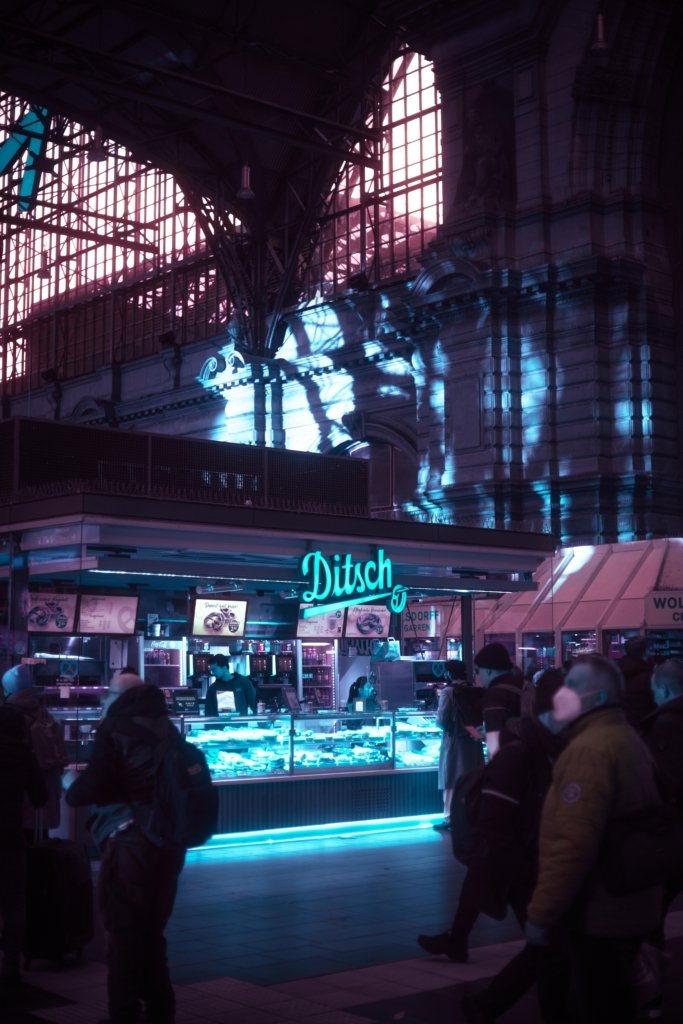 Bäckereistand im Frankfurter Hauptbahnhof bei Nacht mit türkisfarbenem Neonlicht, historische Hallenkonstruktion im Hintergrund, starke Cyberpunk-Farbbearbeitung in Blau und Lila, Menschen im Vordergrund.