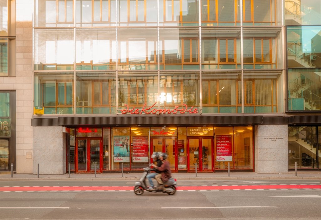Frontansicht der Komödie Frankfurt bei Abendsonne mit goldenem Lichtreflex in der Glasfassade, im Vordergrund ein Paar auf einem Roller in Bewegungsunschärfe.