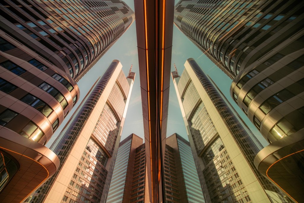 Symmetrische Froschperspektive zwischen den Glastürmen des Frankfurter Bankenviertels, zentrale orangefarbene Neonstreifen als Achse, gewölbte Fassaden links und rechts, türkisfarbener Himmel oben.