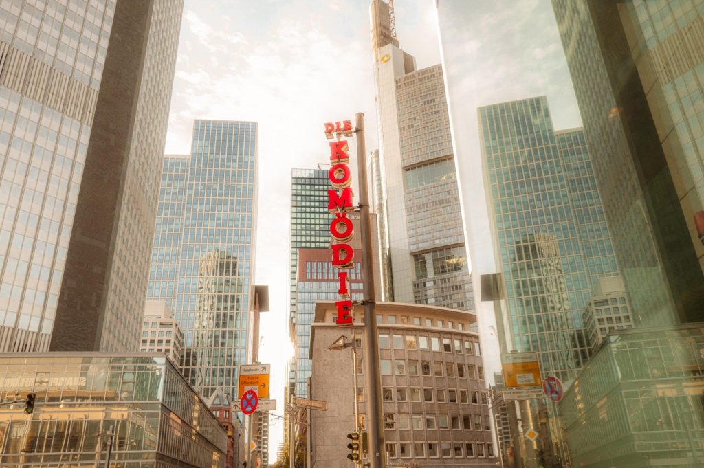 Vertikales rotes Leuchtschild der Komödie Frankfurt von unten fotografiert, im Hintergrund die Glastürme des Frankfurter Bankenviertels bei warmem Gegenlicht.