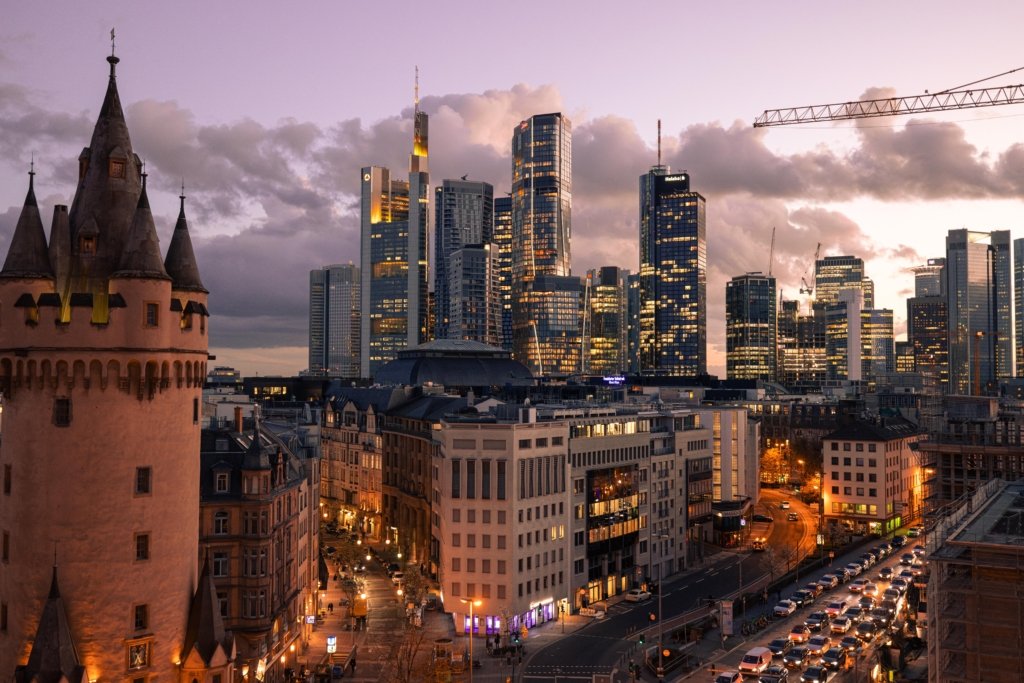 Panoramablick über Frankfurt bei Abenddämmerung, im Vordergrund der mittelalterliche Eschenheimer Turm, im Hintergrund die beleuchtete Skyline des Bankenviertels unter dramatisch bewölktem lila Himmel.