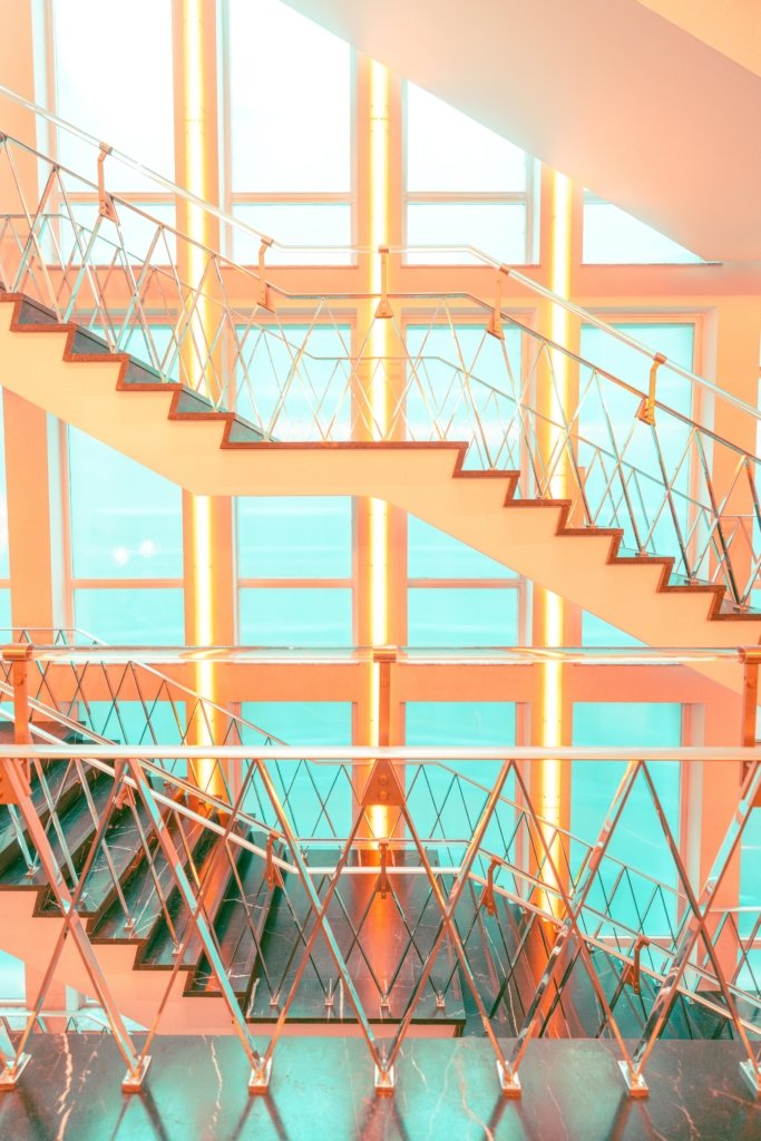 Mehrstöckiges Treppenhaus mit chromfarbenen Geländern in X-Muster-Optik, Farbbearbeitung in Orange und Türkis, große Glasfenster im Hintergrund, Frankfurt.