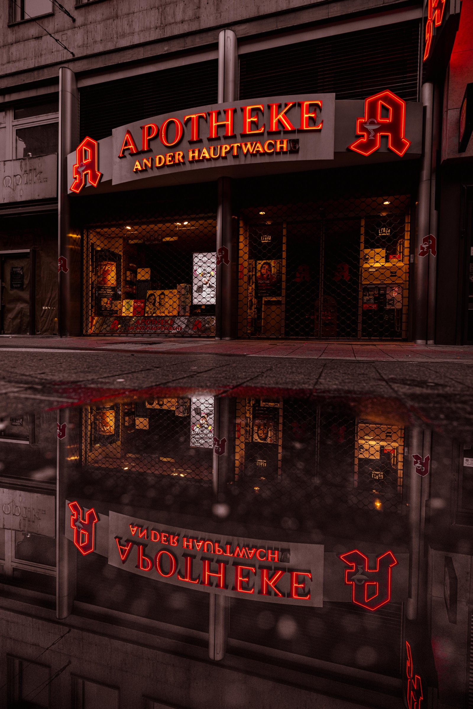 Frankfurt 02.10 16 scaled Rote Neonreklame der Apotheke an der Hauptwache in Frankfurt bei Nacht, perfekt gespiegelt in einer Regenpfütze auf dem Gehweg, geschlossene Sicherheitsgitter sichtbar.