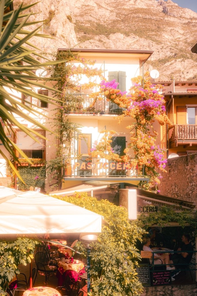 Historisches Gebäude einer Pasticceria in Limone sul Garda, überwuchert von orangefarbener und pinker Bougainvillea, im Vordergrund Palmenblätter und ein Restaurantbereich, im Hintergrund die Felswände über dem Ort.