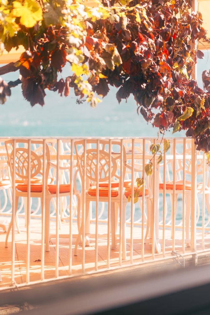 Leere weiße Stühle mit orangefarbenen Kissen auf einer Restaurantterrasse in Limone sul Garda, gerahmt von herbstlich verfärbten Blättern, türkisfarbener Gardasee im Hintergrund.
