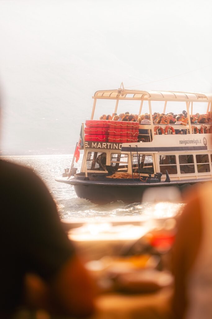 Vollbesetzte Fähre S. Martino auf dem Gardasee bei Gegenlicht, aufgenommen von einem anderen Boot in Limone sul Garda, unscharfe Personen und Getränk im Vordergrund.
