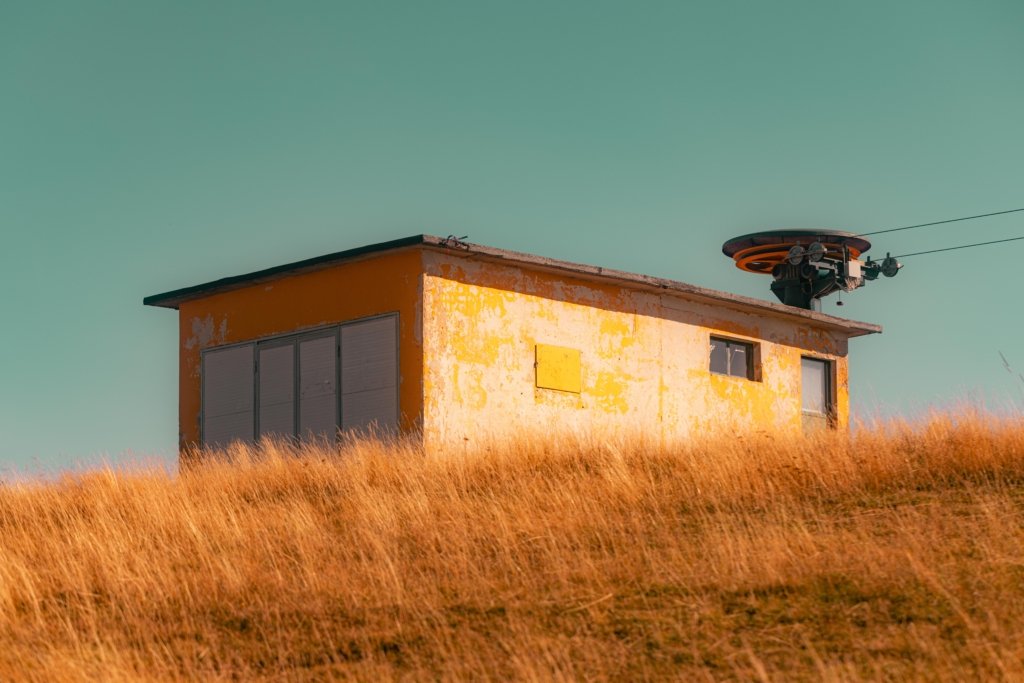 Verlassene verwitterte gelbe Liftstation auf dem Monte Baldo bei Gardasee, umgeben von trockenem goldenen Gras vor türkisblauem Himmel, Seilbahnmechanismus sichtbar.