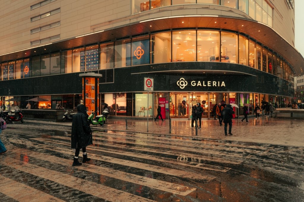 Person auf einem nassen Zebrastreifen auf der Frankfurter Zeil im Winter bei Schneeregen, im Hintergrund die beleuchtete Glasfassade eines Kaufhauses, Straßenreflexionen auf dem Boden.